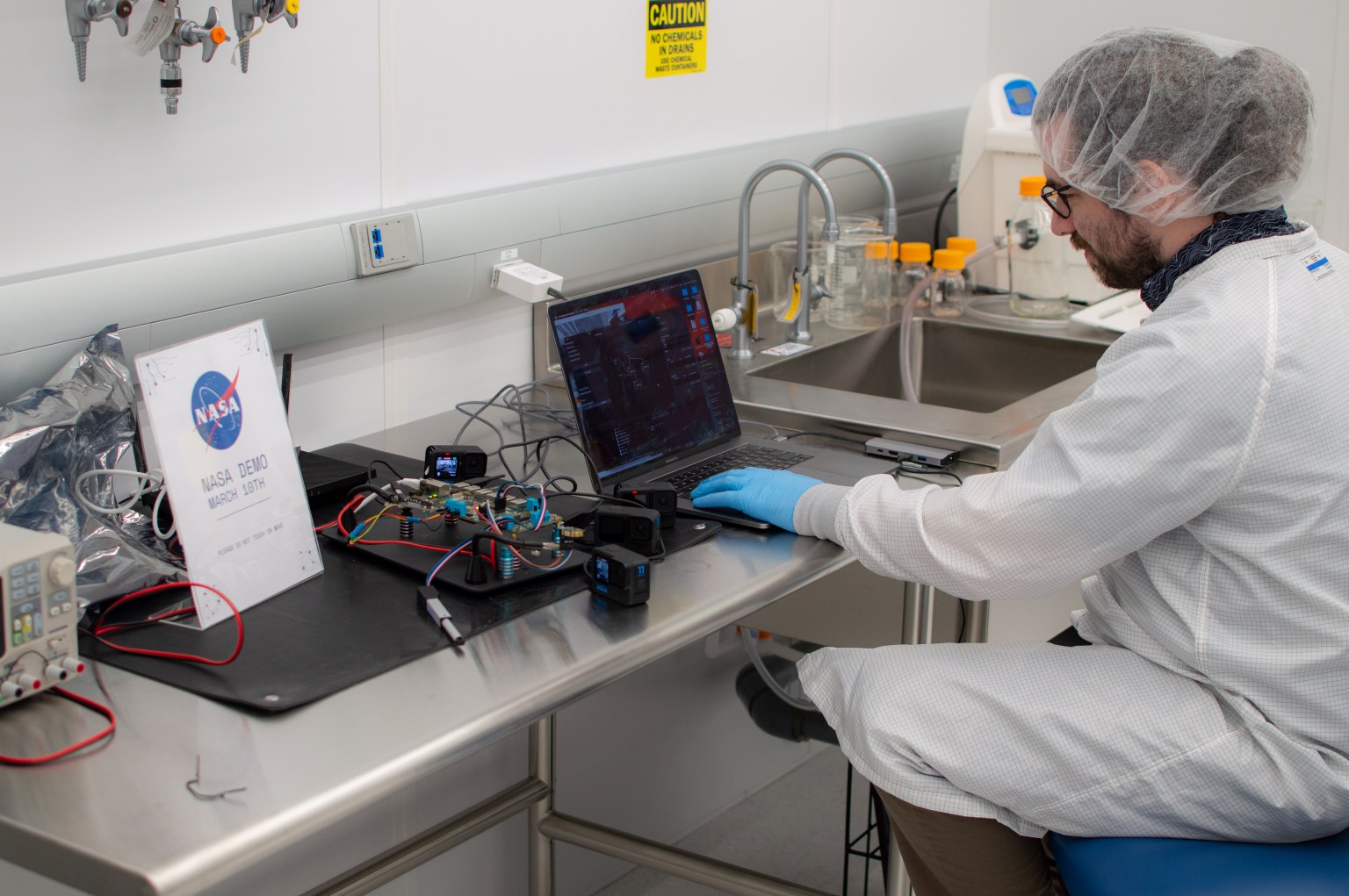 A researcher wearing a white coat, gloves, and hairnet sits at a laptop. A device with multiple colored wires extending from it sits next to him.