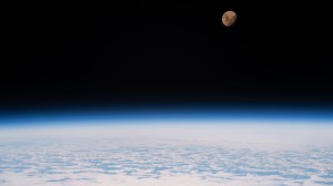 A view of the Moon from the International Space Station as it orbited 264 miles above a partly cloudy Indian Ocean southeast of Madagascar. The Moon has a golden glow to it, appearing more bronze than its usual gray. It stands out against the darkness of space, while below, Earth's atmosphere glows blue. Below that, we can see white clouds covering up most of the water below.