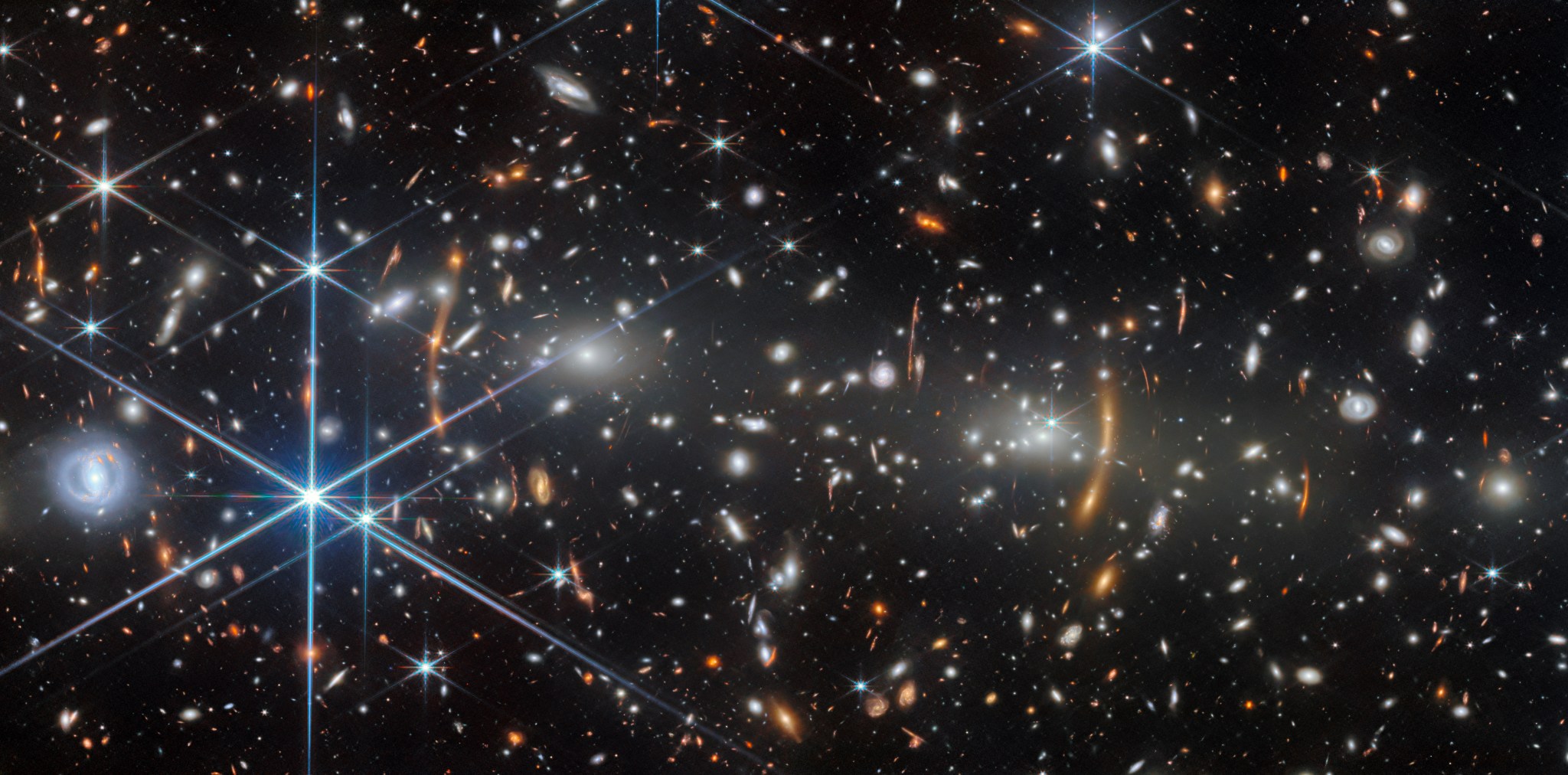 A galaxy cluster in deep space. It is filled with elliptical galaxies: small, bright white glowing ovals. The two largest elliptical galaxies, left and right of center, are bright cores that radiate light. Unrelated, distant galaxies are scattered around as red smudges and dots.Many of these are stretched out into red arcs and lines by the galaxy cluster’s strong gravity, creating multiple images in places. Numerous spiral galaxies and bright stars appear in the foreground.