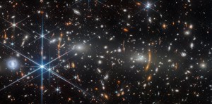 A galaxy cluster in deep space. It is filled with elliptical galaxies: small, bright white glowing ovals. The two largest elliptical galaxies, left and right of center, are bright cores that radiate light. Unrelated, distant galaxies are scattered around as red smudges and dots.Many of these are stretched out into red arcs and lines by the galaxy cluster’s strong gravity, creating multiple images in places. Numerous spiral galaxies and bright stars appear in the foreground.