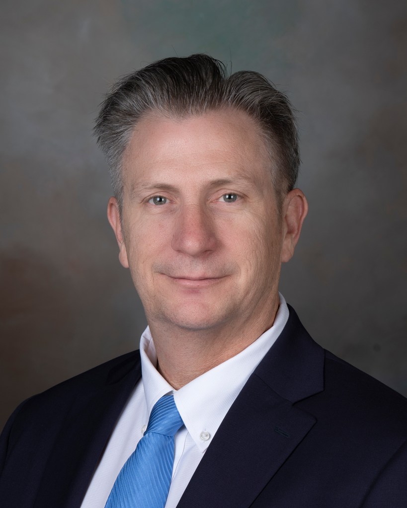 Portrait of a man wearing a black suit jacked, blue tie, and white shirt.