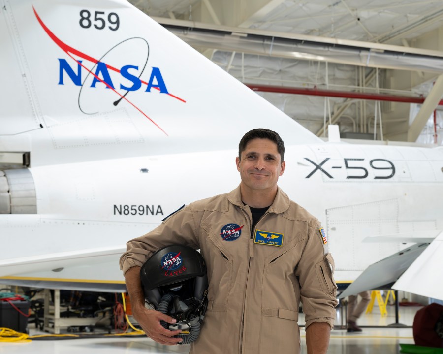NASA’s X-59 test pilot Mike Lippert stands in a hangar in front of NASA’s quiet supersonic research aircraft, wearing a tan NASA flight suit and holding a flight helmet under the right arm. The X-plane in the background displays the NASA logo, tail number 859, and “X-59” marking.