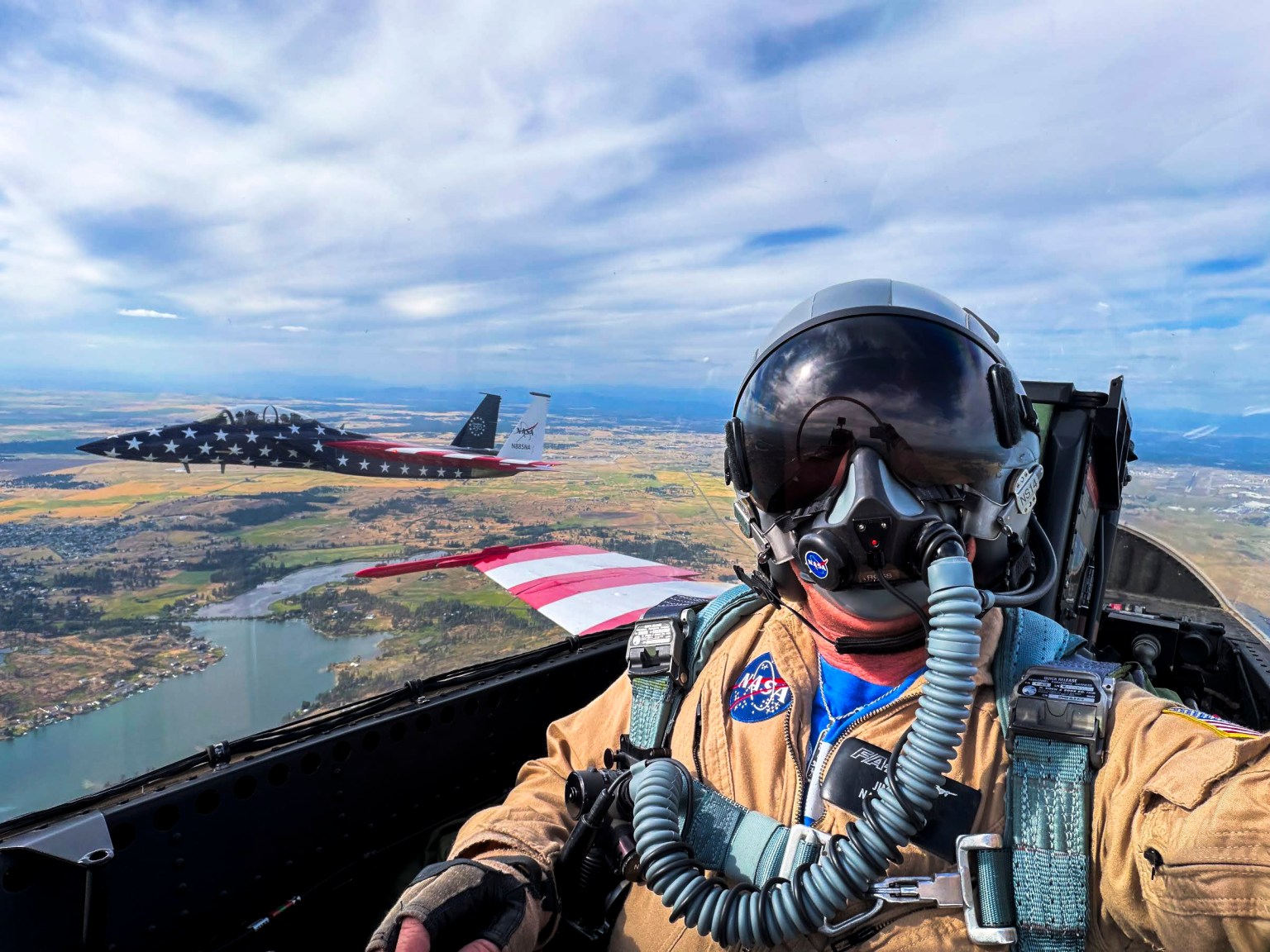 A photographer takes a selfie from the rear seat of a jet aircraft during flight, with another jet visible through the window. Each jet is painted in the United States flag.