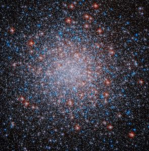A packed, roughly spherical cluster of red, white and blue stars shines against the black background of space. The stars are clustered most densely toward the center of the cluster.