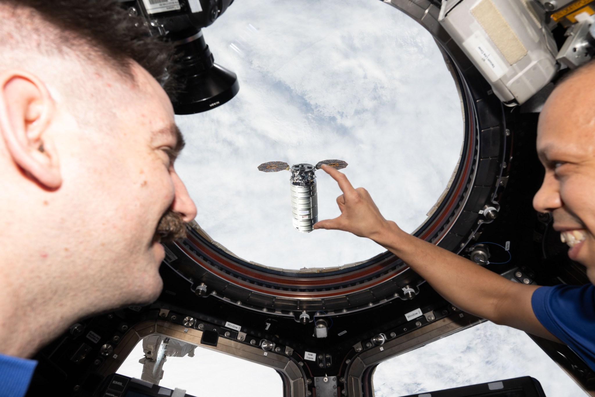 NASA astronauts Jack Hathaway (left) and Chris Williams (right) look through a circular window in the International Space Station's cupola as Northrop Grumman’s Cygnus XL spacecraft approaches. The spacecraft is a silver cylinder with two hexagonal solar arrays extending from either side of the top end of the cylinder. Behind the spacecraft, white clouds cover the Earth.