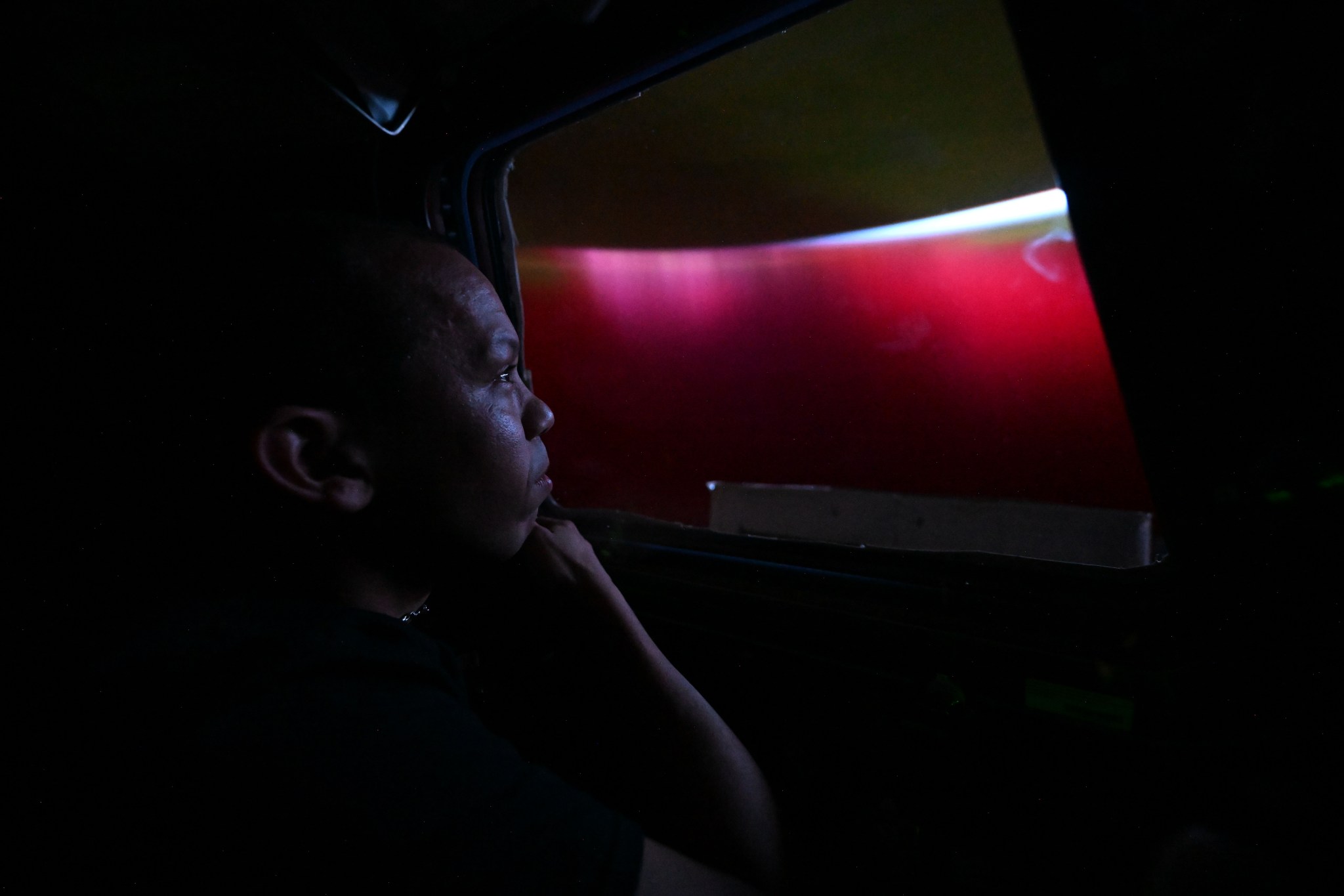 NASA astronaut Chris Williams gazes out of a trapezoid-shaped window at a glowing red aurora above the Earth. The curvature of the Earth is visible through a bright green curve. It is dark aboard the International Space Station in the photo, and the glow from the aurora softly illuminates Williams' face.