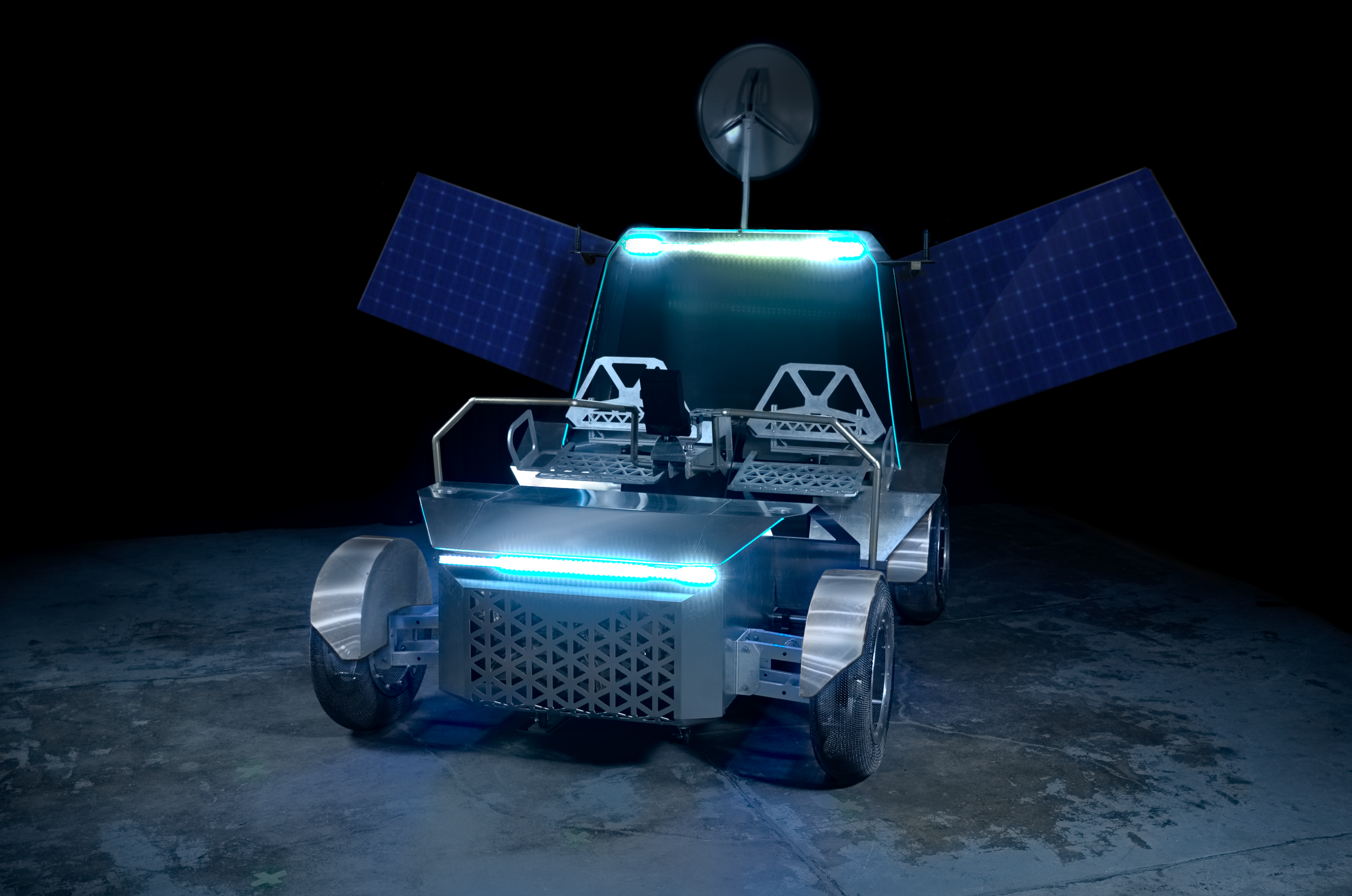 A photo of a full-scale prototype of Lunar Outpost's Pegasus lunar terrain vehicle. The rover can seat two astronauts, is fabricated out of metal, has mesh tires, has two solar arrays deployed, and is lit up by blue lights.