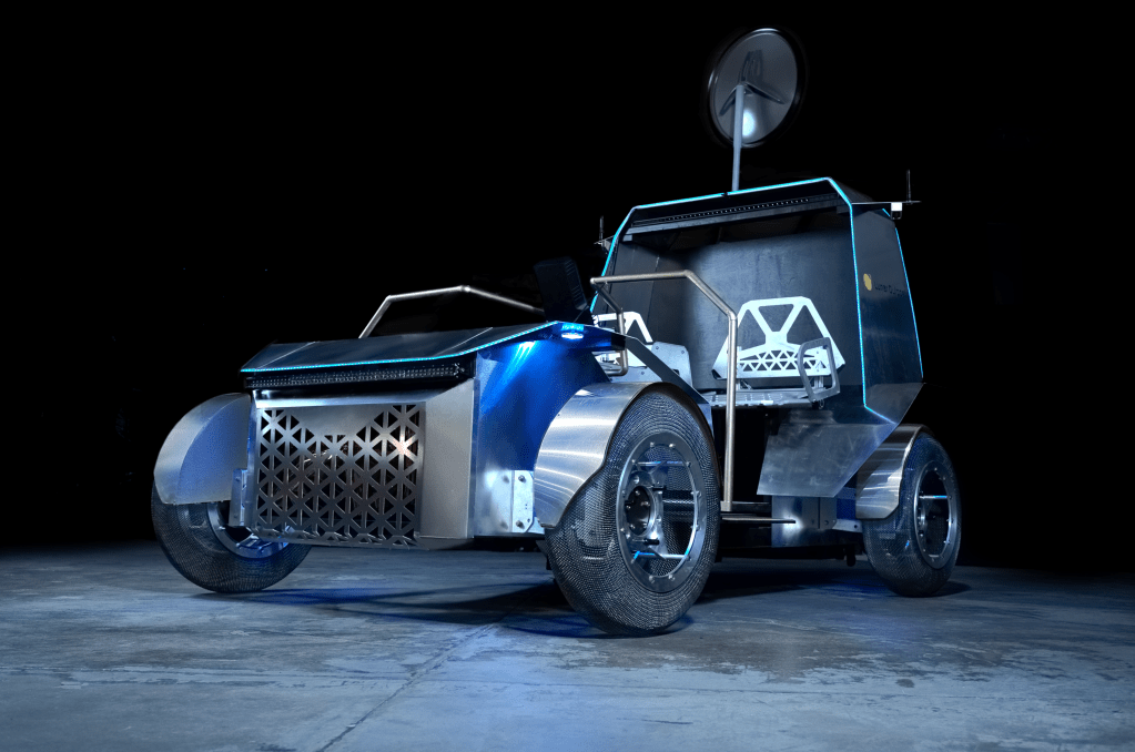 A photo of a full-scale prototype of Lunar Outpost's Pegasus lunar terrain vehicle. The rover can seat two astronauts, is fabricated out of metal, has mesh tires, and is lit up by blue lights.