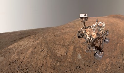 A wide-angle, color selfie of NASA's Perseverance rover on a sloping, reddish-brown Martian landscape. The rover is positioned on the right side of the image, showing its intricate metallic body, six treaded wheels, and tall camera mast, which is turned to "look" directly at the viewer. The rocky, textured ground slopes downward to the left, revealing a vast, desolate basin and distant crater rims under a dusty, pale grayish-tan sky.
