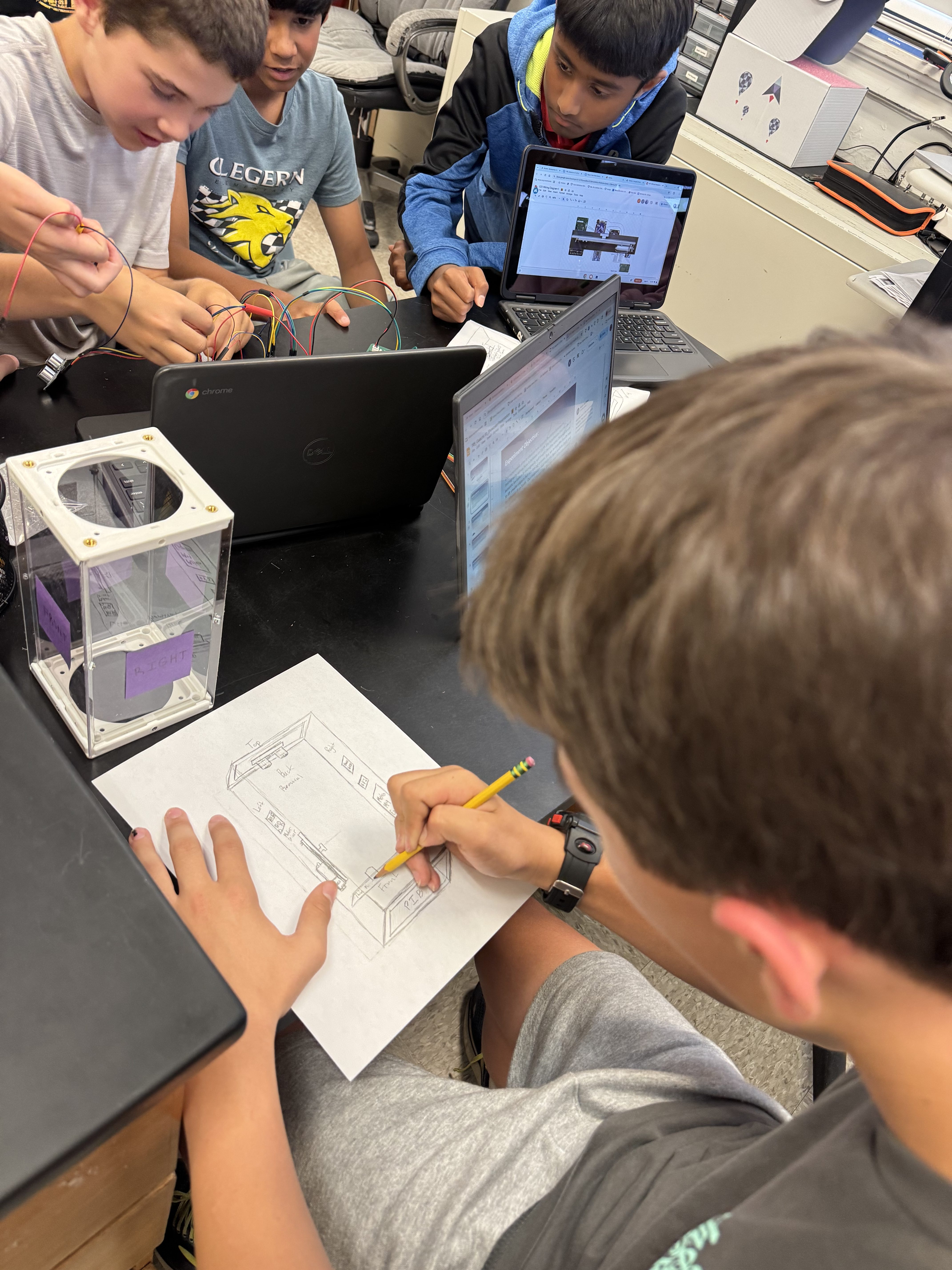 Several students site at a table. One student is drawing a wiring diagram.