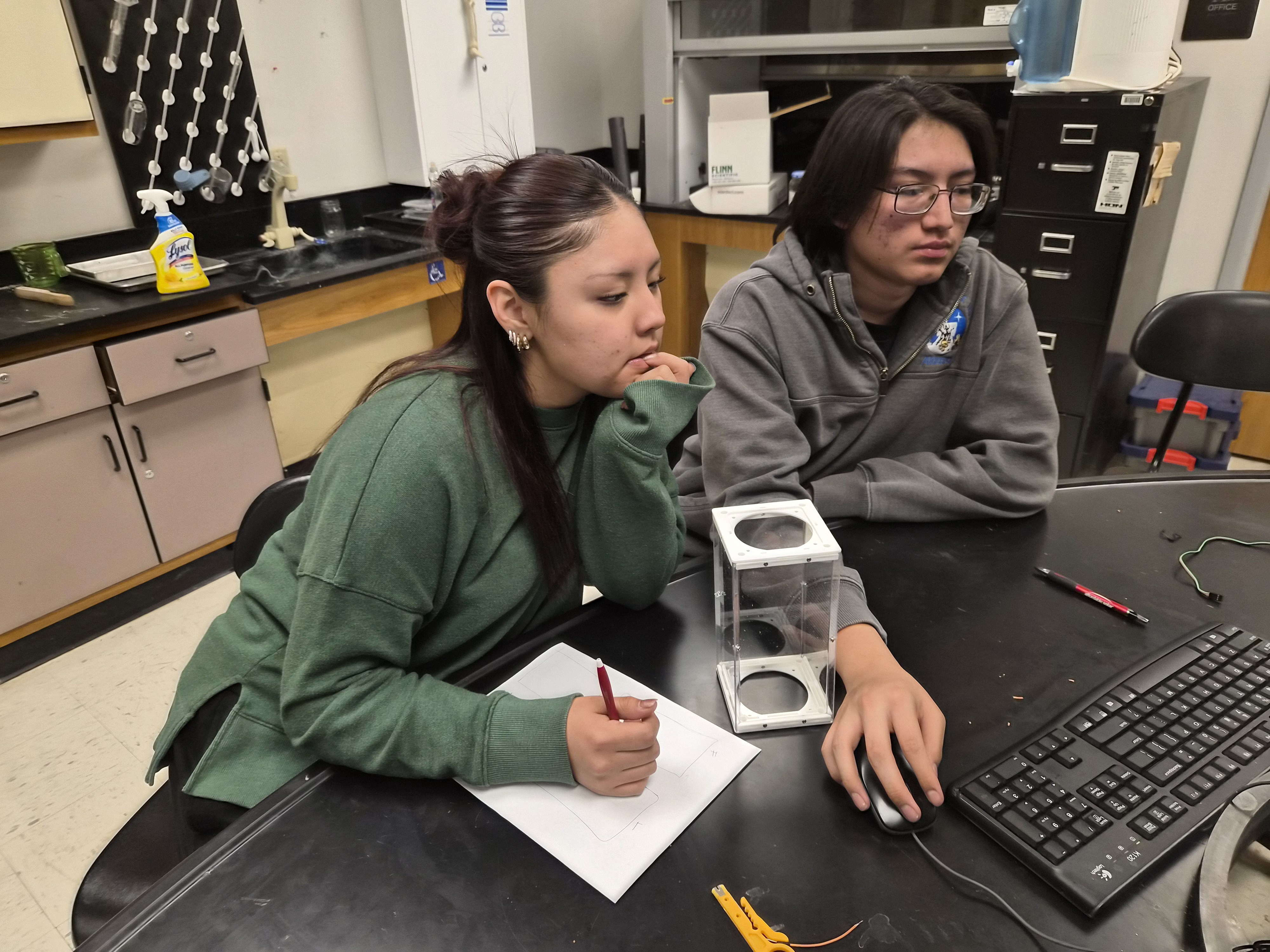 Two students sit at a table with a laptop. A cube-shaped container sits in front of them.