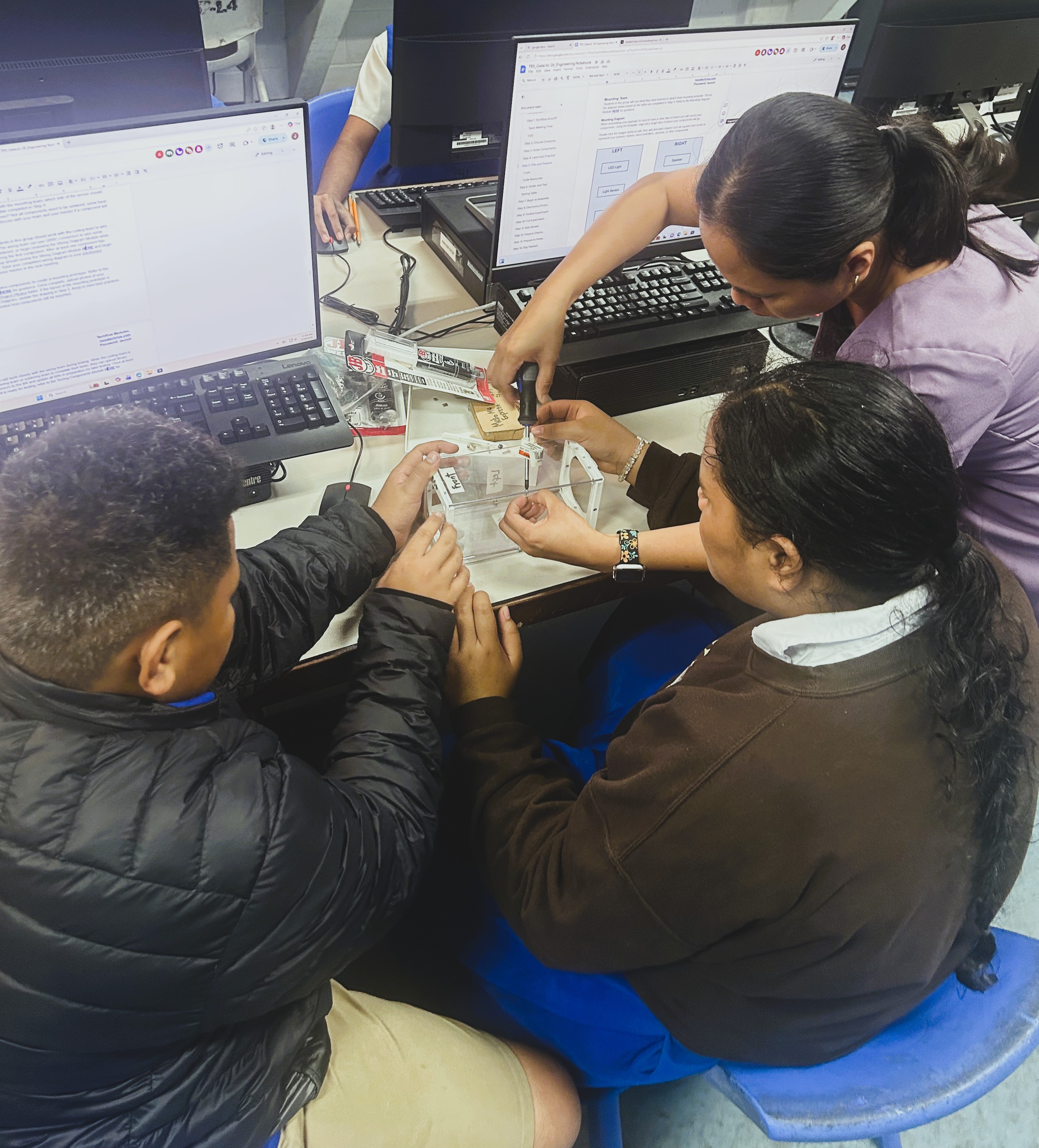 Three students viewed from behind. Two are holding a clear acrylic box while a third student uses a screwdriver to attach a component.