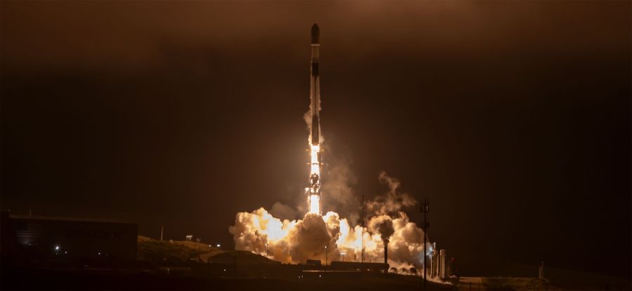 A rocket launches at night, producing bright flames and smoke as it lifts off from a dimly lit launchpad.