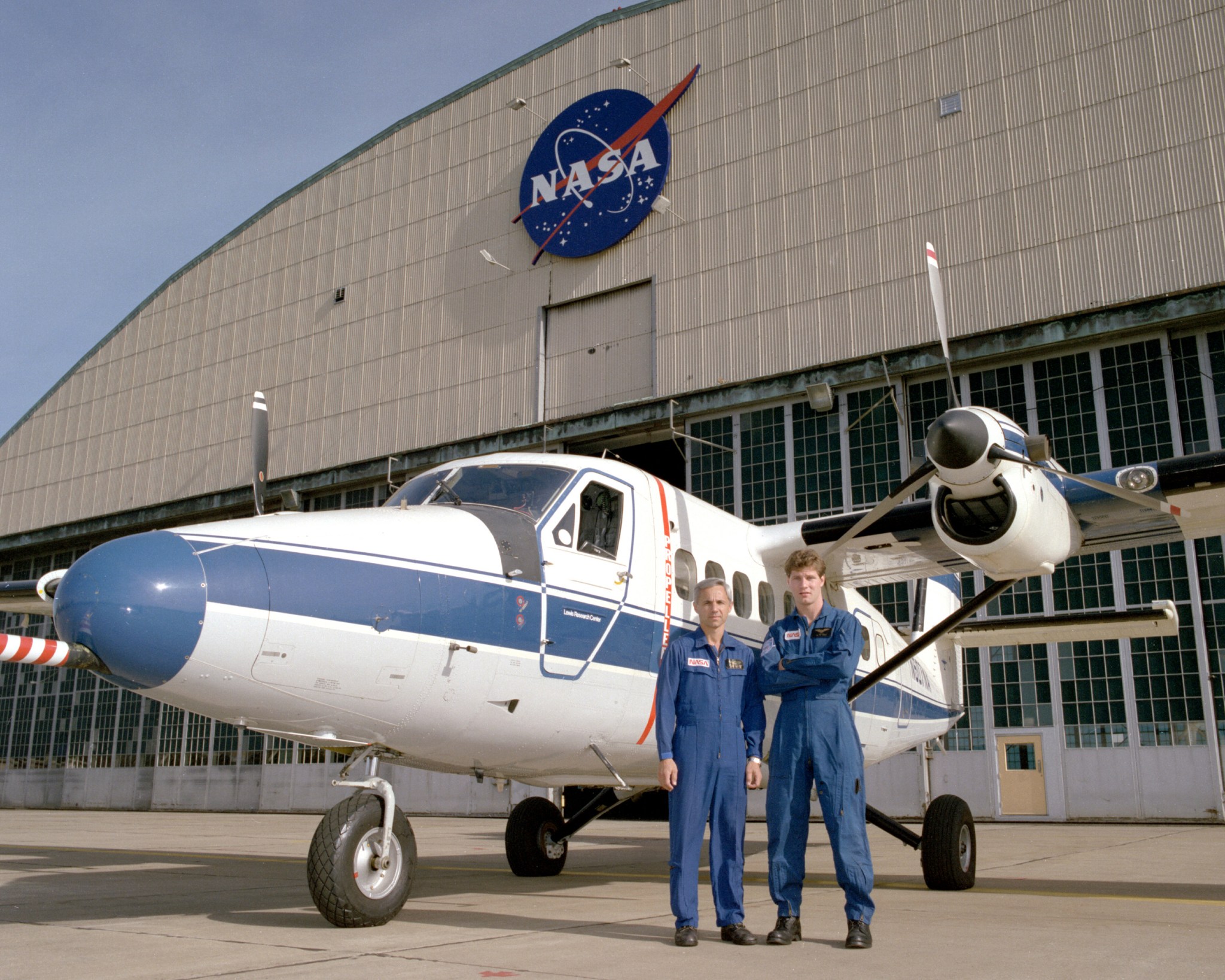 Two men in flight suits stand in front of a small aircraft painted white and blue. Behind it is an aircraft hangar with the NASA logo at the top.