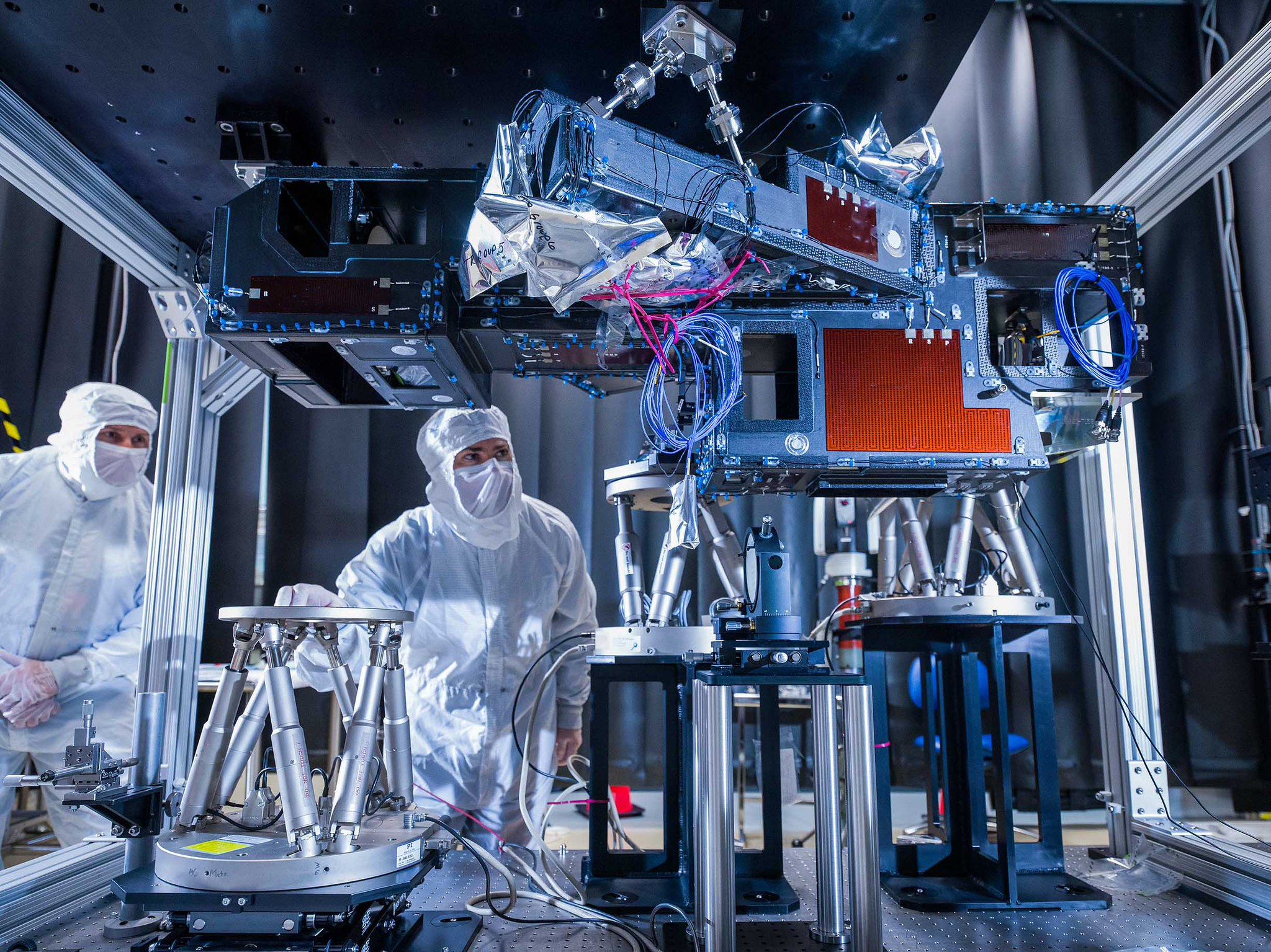 Engineers at L3Harris Technologies, a contractor for the Roman Coronagraph Instrument, are shown inspecting the instrument’s optical path, which feeds light from the telescope into the instrument. Two people on the left of the image are wearing white full-body suits and are bent down, looking up at the instrument components. The hardware in front of them are made up of metal boxes and electrical wires, set on pedestals that lift them up above the engineers’ heads. The topmost instrument has textured red sides and blue and pink wires. All of the components are inside a silver framework with a black panel on top. Photo credit: Chris Gunn/ NASA JPL