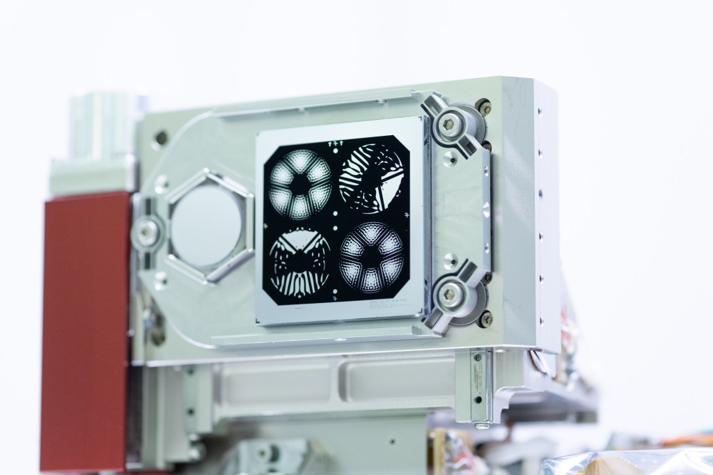 Close-up shot of four of the coronagraph’s masks. The masks are black shapes painted on a reflective silver square. The square is mounted on a metal rectangular box. The the top left and bottom right masks look like a flowers with six petals; the top right and bottom left masks look like insects with their wings spread open.