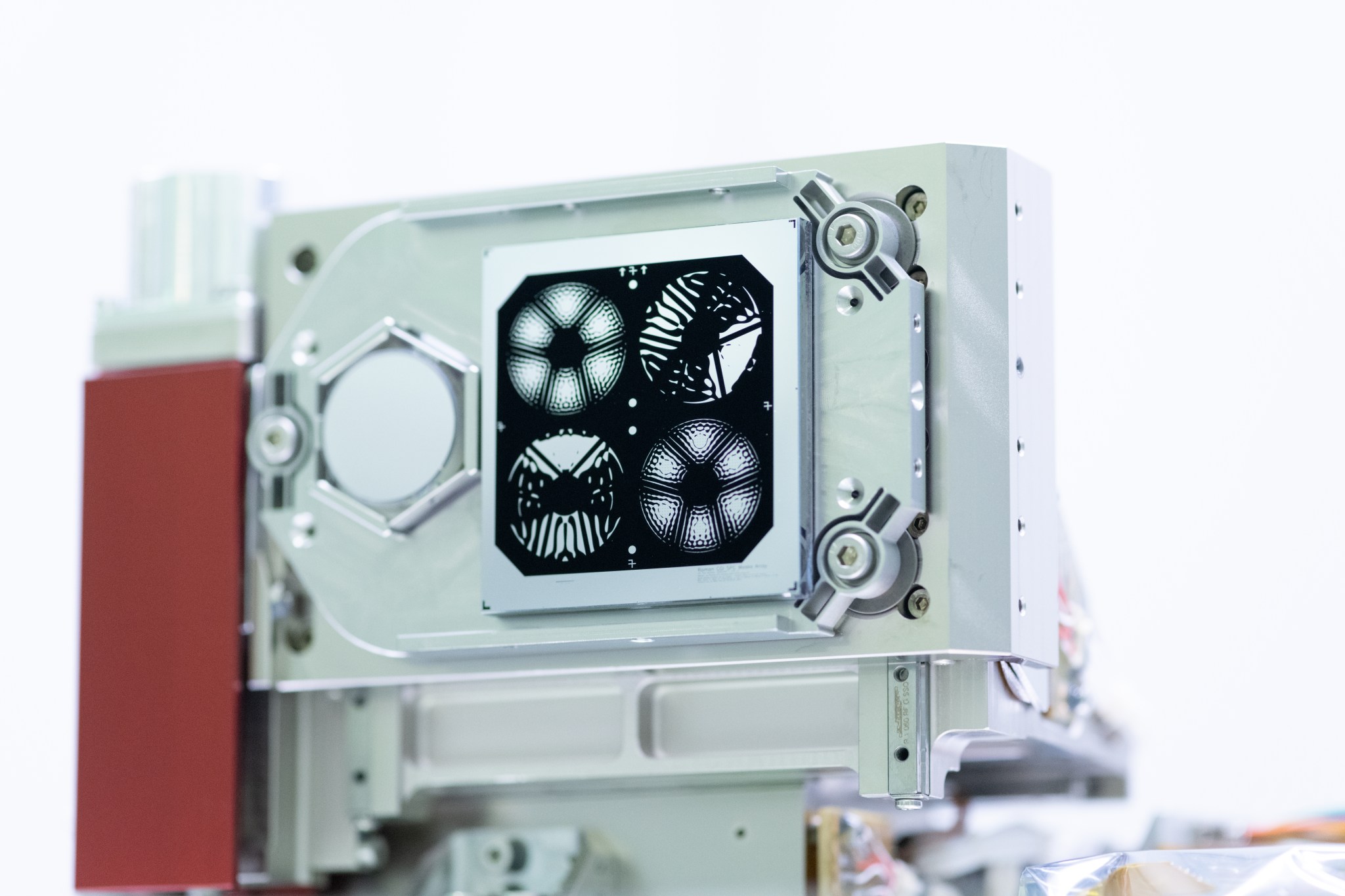 Close-up shot of four of the coronagraph’s masks. The masks are black shapes painted on a reflective silver square. The square is mounted on a metal rectangular box. The the top left and bottom right masks look like a flowers with six petals; the top right and bottom left masks look like insects with their wings spread open.