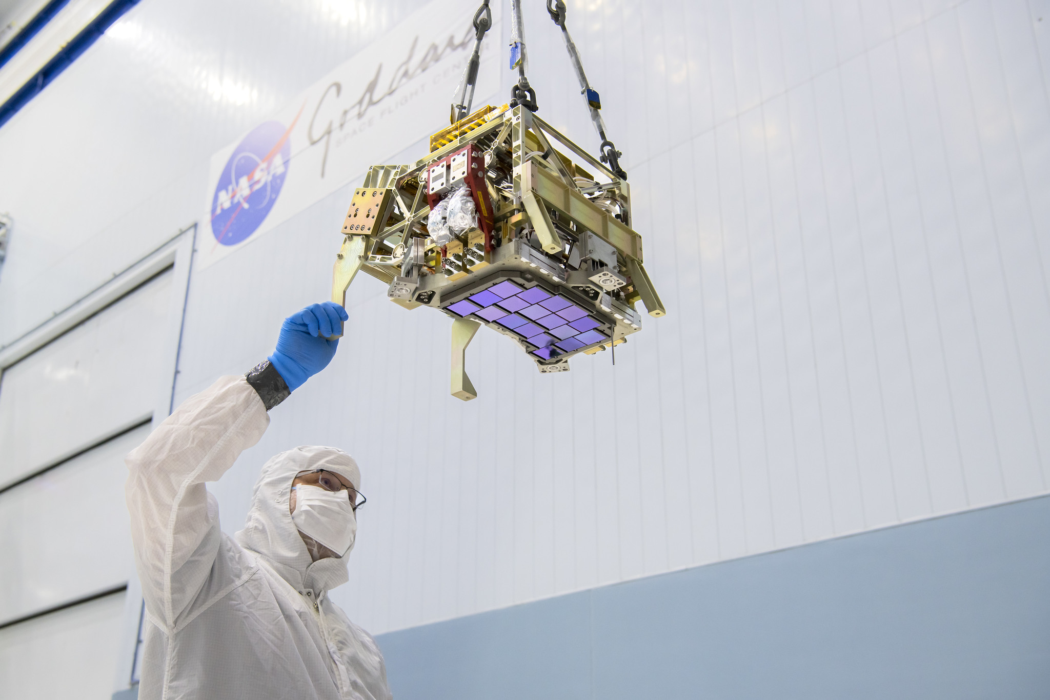 An iridescent purplish-blue array of square detectors attached to a chunky, rectangular piece of gold and silver metal hardware is seen from below. A man in a white suit and gloves holds onto an edge of the structure. The structure hangs from the ceiling of a large room with white and blue walls. It is about the size of a small microwave, and the array of detectors is arranged in slightly curved rows. Out of focus in the background, the blue and red NASA logo and text “Goddard Space Flight Center” are visible. Credits: NASA/Chris Gunn