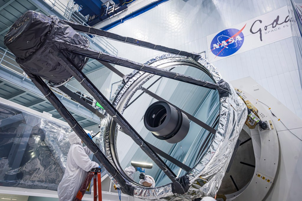 An engineer wearing a baggy white clean room gown inspects the Roman Space Telescope's primary mirror. The mirror is a large reflective circle embedded in other large machinery.