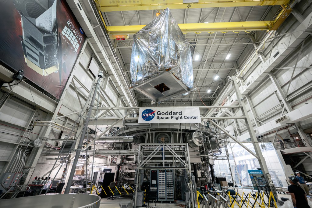 A portion of the Roman Space Telescope inside a silver protective cover is suspended above the Space Environment Simulator, a huge stainless steel chamber.