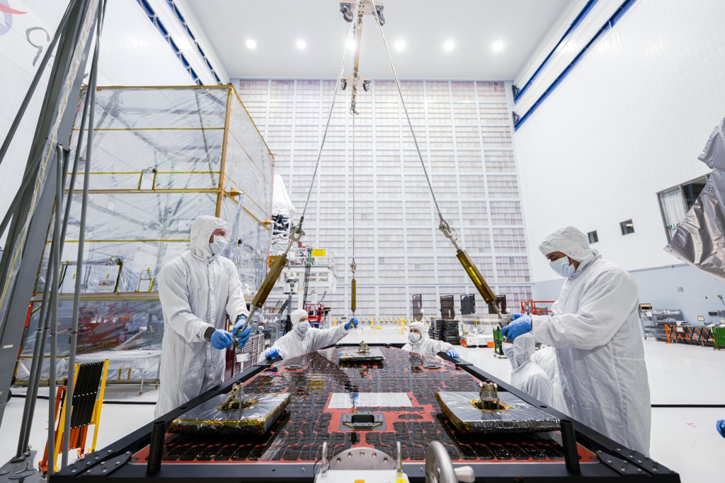 Four technicians work on hardware for the Roman Space Telescope. The technicians are all wearing white coveralls, including face masks and gloves. They are working on Roman's solar panels, which are orange and black, angled horizontally, parallel to the ground. The technicians are inside the huge clean room at NASA's Goddard Space Flight Center, and a large grid of air filters is visible on the wall behind them.