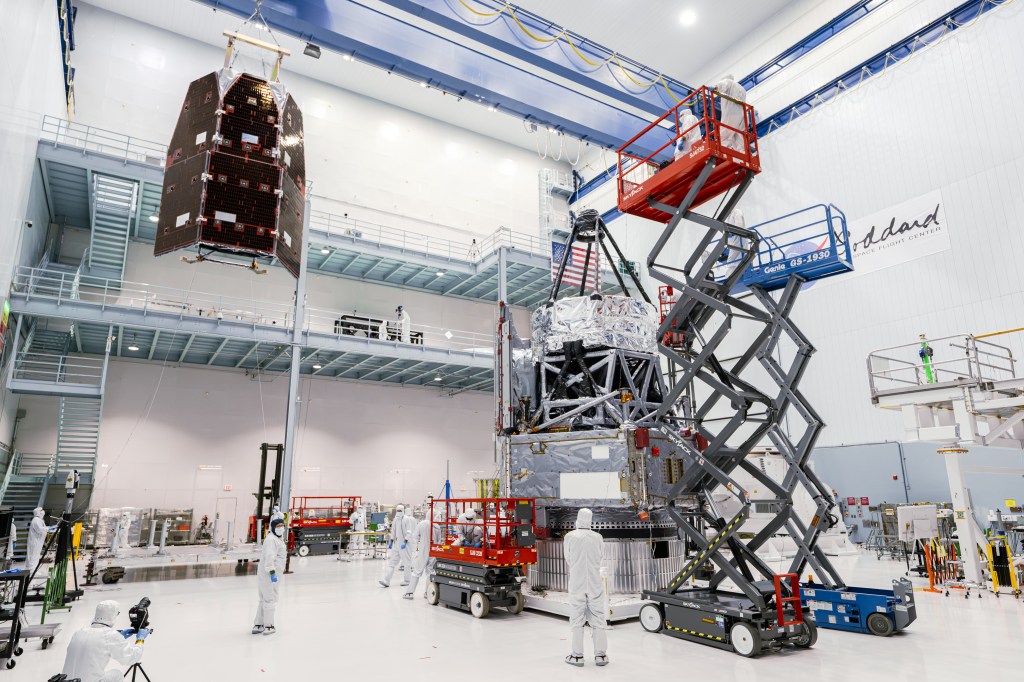 Several technicians -- all wearing white coveralls inside a huge clean room -- work on the Roman Space Telescope. Some technicians stand on the ground, and some are high above the ground using heavy machinery.
