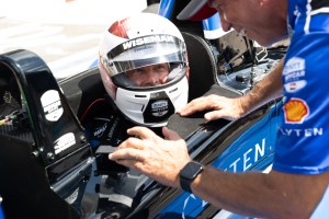 NASA astronaut Reid Wiseman looks up at another person speaking. He is sitting in the second seat of a racecar. He's wearing a white helmet with his last name on the visor.