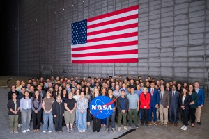 A large group of people pose for a picture inside a massive wind tunnel. There is a large American flag on the wall behind them. A few people in the front hold up the NASA "meatball" logo.