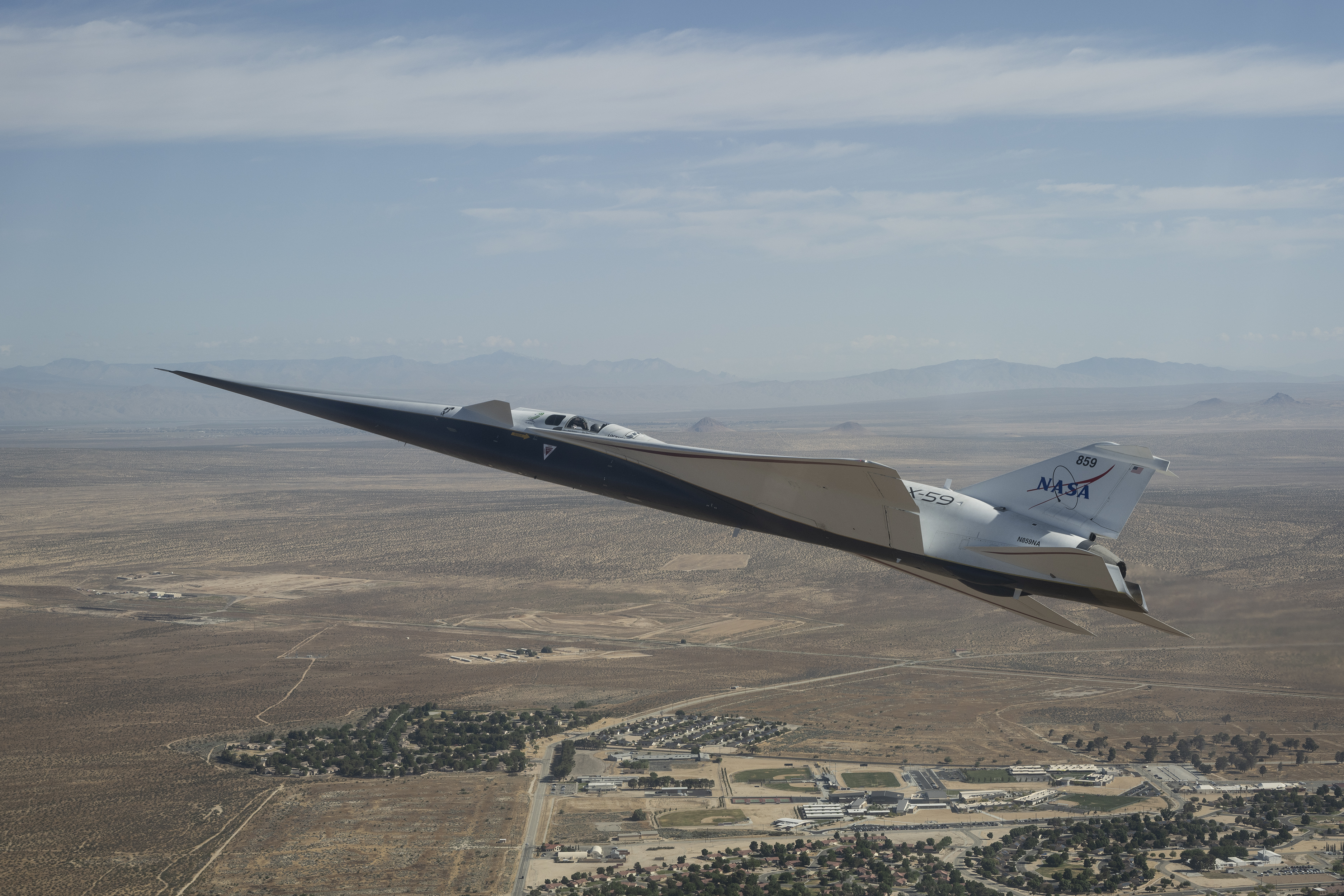 NASA’s white and blue X-59 quiet supersonic research aircraft is shown flying above the desert near NASA’s Armstrong Flight Research Center in California. Viewed from the side, the aircraft’s long nose, swept wings and narrow fuselage stand out against the desert landscape.
