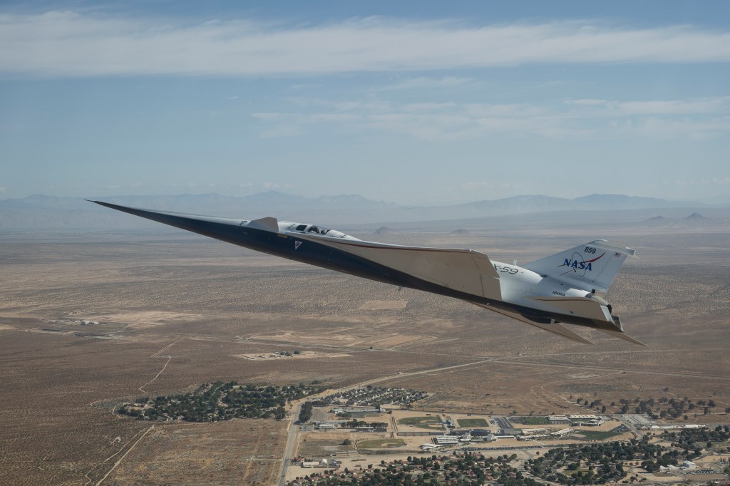 NASA’s white and blue X-59 quiet supersonic research aircraft is shown flying above the desert near NASA’s Armstrong Flight Research Center in California. Viewed from the side, the aircraft’s long nose, swept wings and narrow fuselage stand out against the desert landscape.