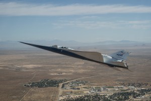 NASA’s white and blue X-59 quiet supersonic research aircraft is shown flying above the desert near NASA’s Armstrong Flight Research Center in California. Viewed from the side, the aircraft’s long nose, swept wings and narrow fuselage stand out against the desert landscape.