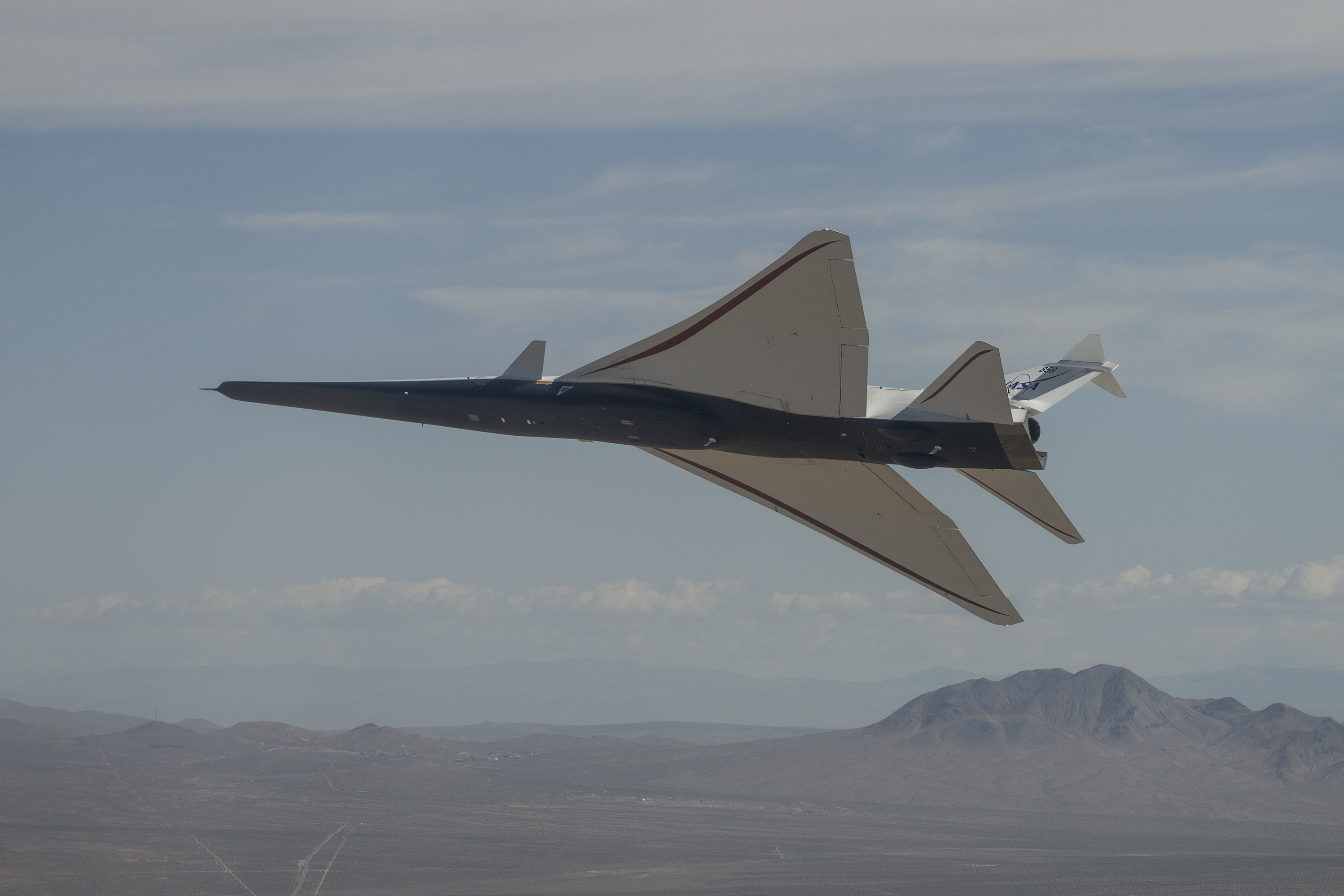 NASA’s X-59 quiet supersonic research aircraft banks above the California desert during a test flight. Viewed from below, the aircraft’s long nose, triangular wings and underside are shown against the desert terrain.