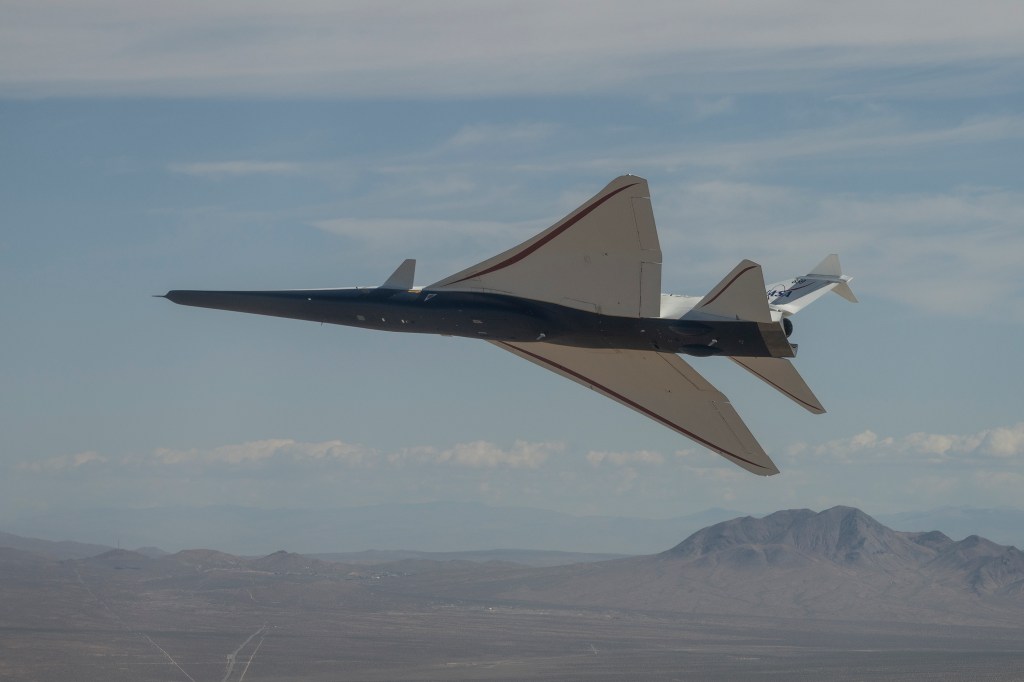 NASA’s X-59 quiet supersonic research aircraft banks above the California desert during a test flight. Viewed from below, the aircraft’s long nose, triangular wings and underside are shown against the desert terrain.