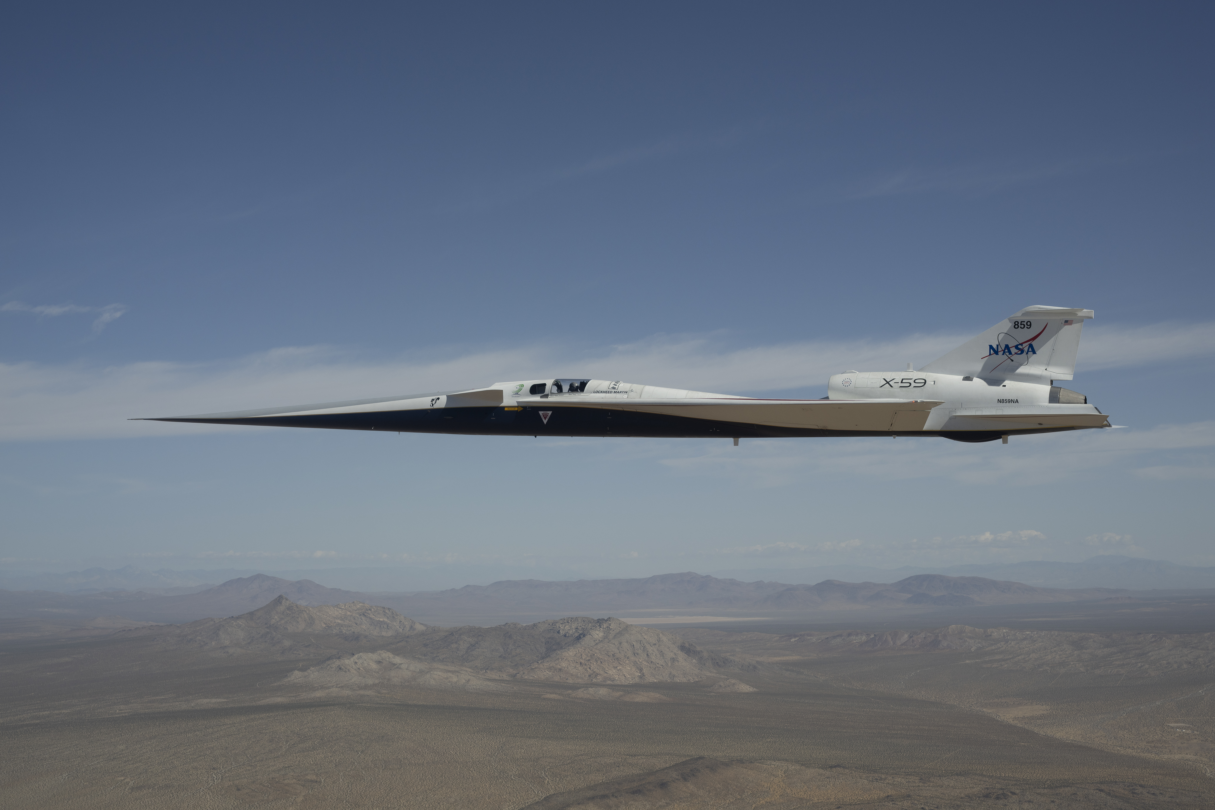 NASA’s white and blue X-59 quiet supersonic research aircraft is shown flying above the desert near NASA’s Armstrong Flight Research Center in California. Viewed from the side, the aircraft’s long nose, swept wings and narrow fuselage stand out against the desert landscape.
