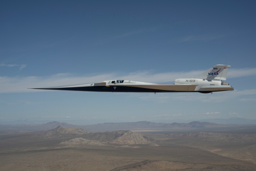 NASA’s white and blue X-59 quiet supersonic research aircraft is shown flying above the desert near NASA’s Armstrong Flight Research Center in California. Viewed from the side, the aircraft’s long nose, swept wings and narrow fuselage stand out against the desert landscape.