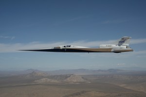 NASA’s white and blue X-59 quiet supersonic research aircraft is shown flying above the desert near NASA’s Armstrong Flight Research Center in California. Viewed from the side, the aircraft’s long nose, swept wings and narrow fuselage stand out against the desert landscape.