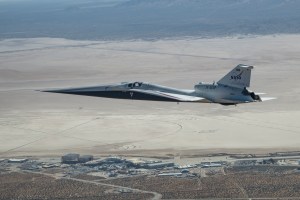 NASA’s X-59 quiet supersonic research aircraft flies above NASA’s Armstrong Flight Research Center and Rogers Dry Lake in California. The aircraft is shown in side profile during level flight with the compass rose, facilities, and desert terrain below.
