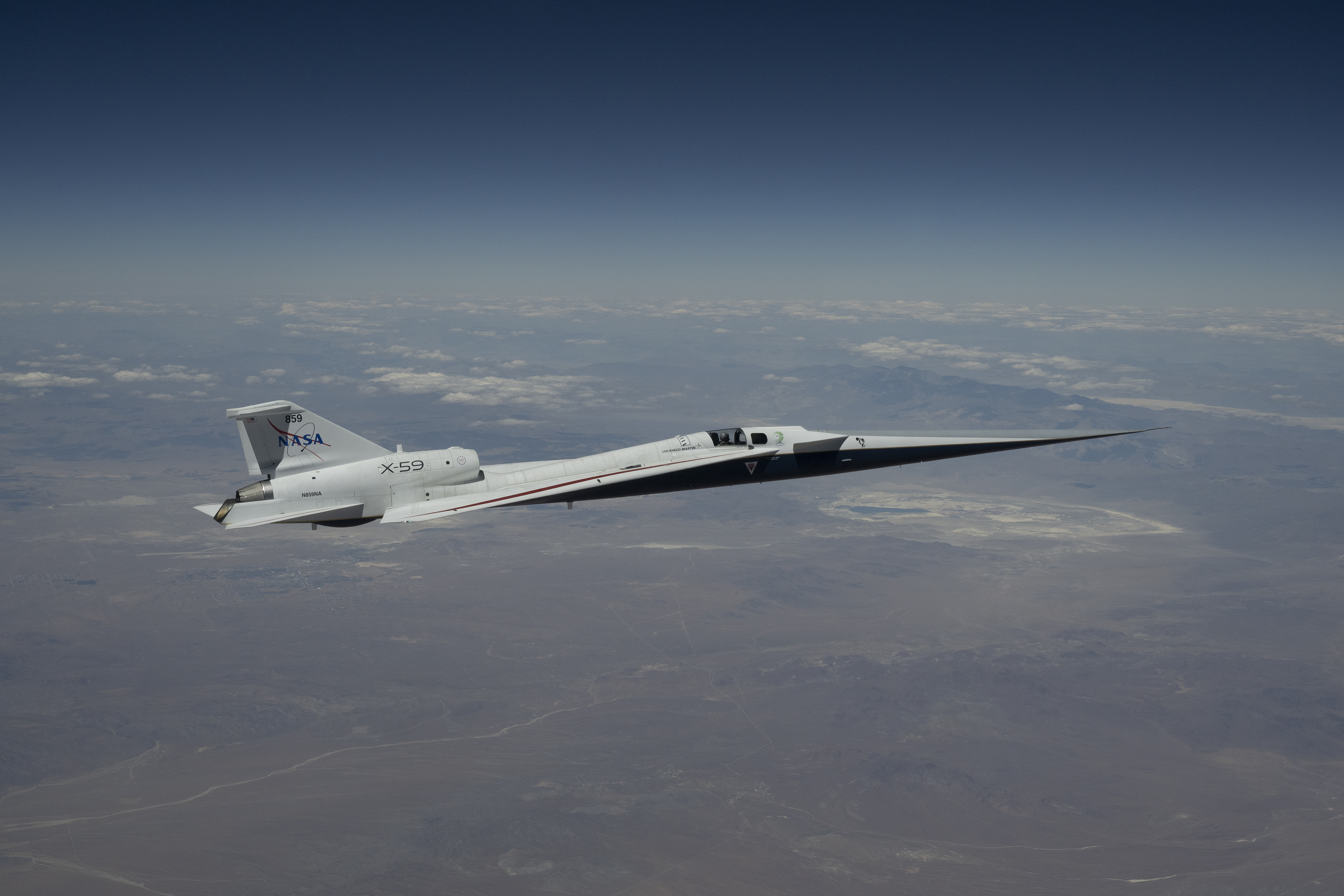 NASA’s X-59 quiet supersonic research aircraft flies high above the Mojave Desert. The aircraft is shown from a side profile with its long nose, swept wings and white fuselage stand out against the background.