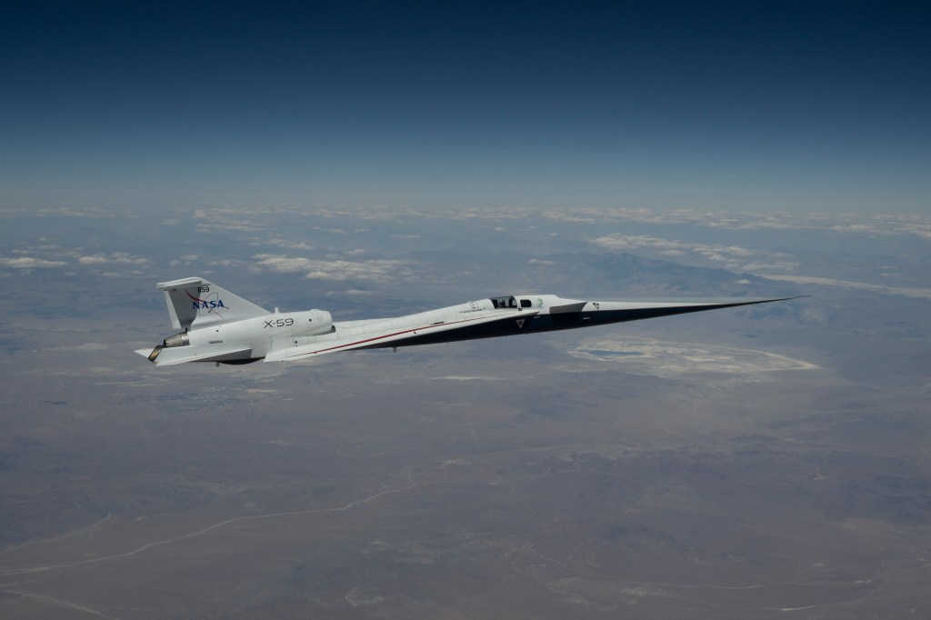 NASA’s X-59 quiet supersonic research aircraft flies high above the Mojave Desert. The aircraft is shown from a side profile with its long nose, swept wings and white fuselage stand out against the background.