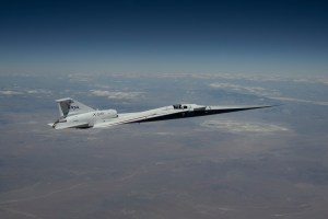 NASA’s X-59 quiet supersonic research aircraft flies high above the Mojave Desert. The aircraft is shown from a side profile with its long nose, swept wings and white fuselage stand out against the background.