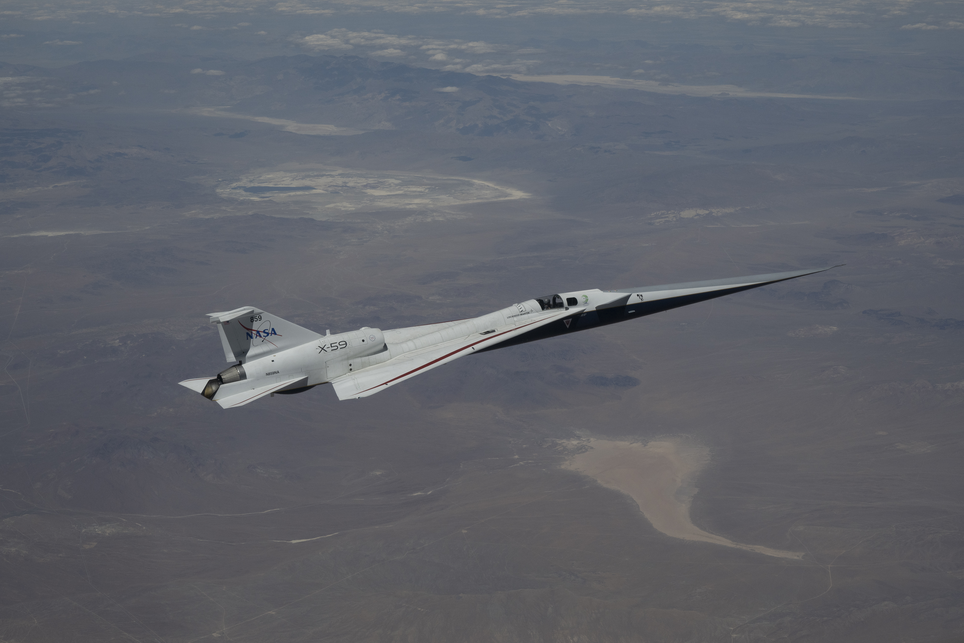 NASA’s X-59 quiet supersonic research aircraft flies high above the Mojave Desert. The aircraft is shown from a side profile with its long nose, swept wings and white fuselage stand out against the background.