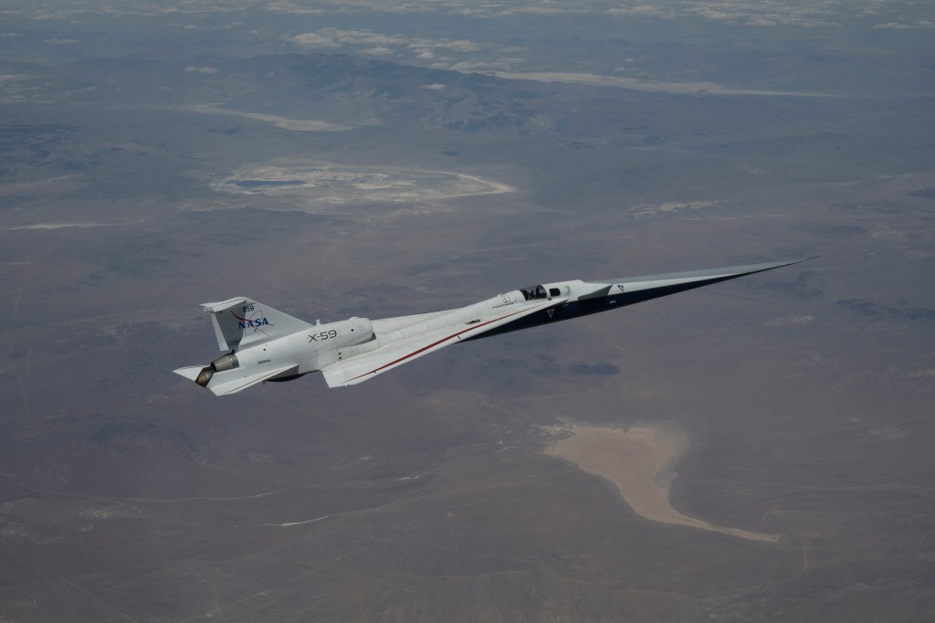 NASA’s X-59 quiet supersonic research aircraft flies high above the Mojave Desert. The aircraft is shown from a side profile with its long nose, swept wings and white fuselage stand out against the background.