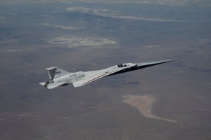 NASA’s X-59 quiet supersonic research aircraft flies high above the Mojave Desert. The aircraft is shown from a side profile with its long nose, swept wings and white fuselage stand out against the background.