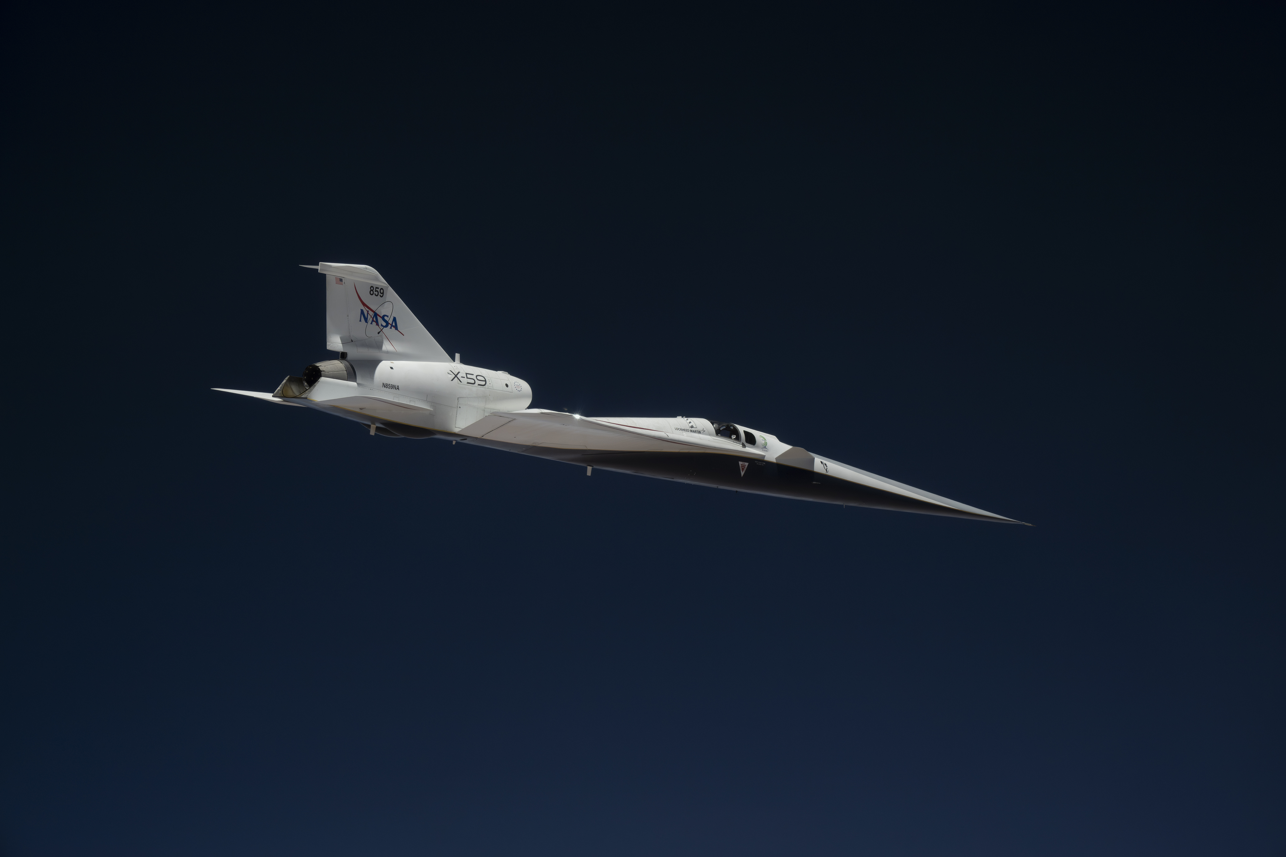 NASA’s X-59 quiet supersonic research aircraft flies high above the Mojave Desert. The aircraft is shown from a side profile with its long nose, swept wings and white fuselage stand out against the background.