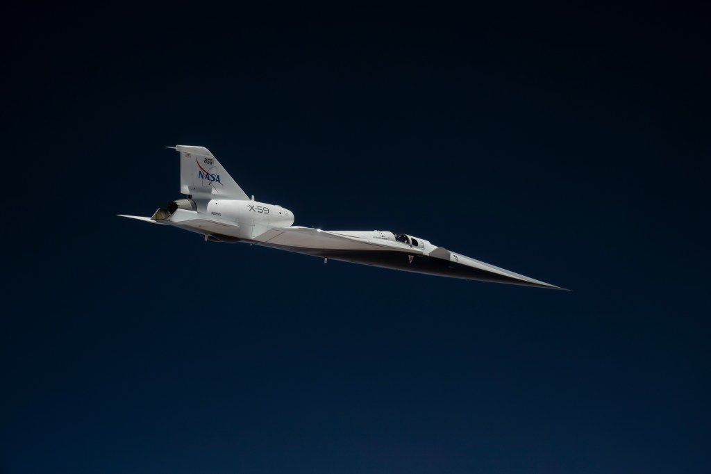 NASA’s X-59 quiet supersonic research aircraft flies high above the Mojave Desert. The aircraft is shown from a side profile with its long nose, swept wings and white fuselage stand out against the background.