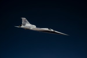 NASA’s X-59 quiet supersonic research aircraft flies high above the Mojave Desert. The aircraft is shown from a side profile with its long nose, swept wings and white fuselage stand out against the background.