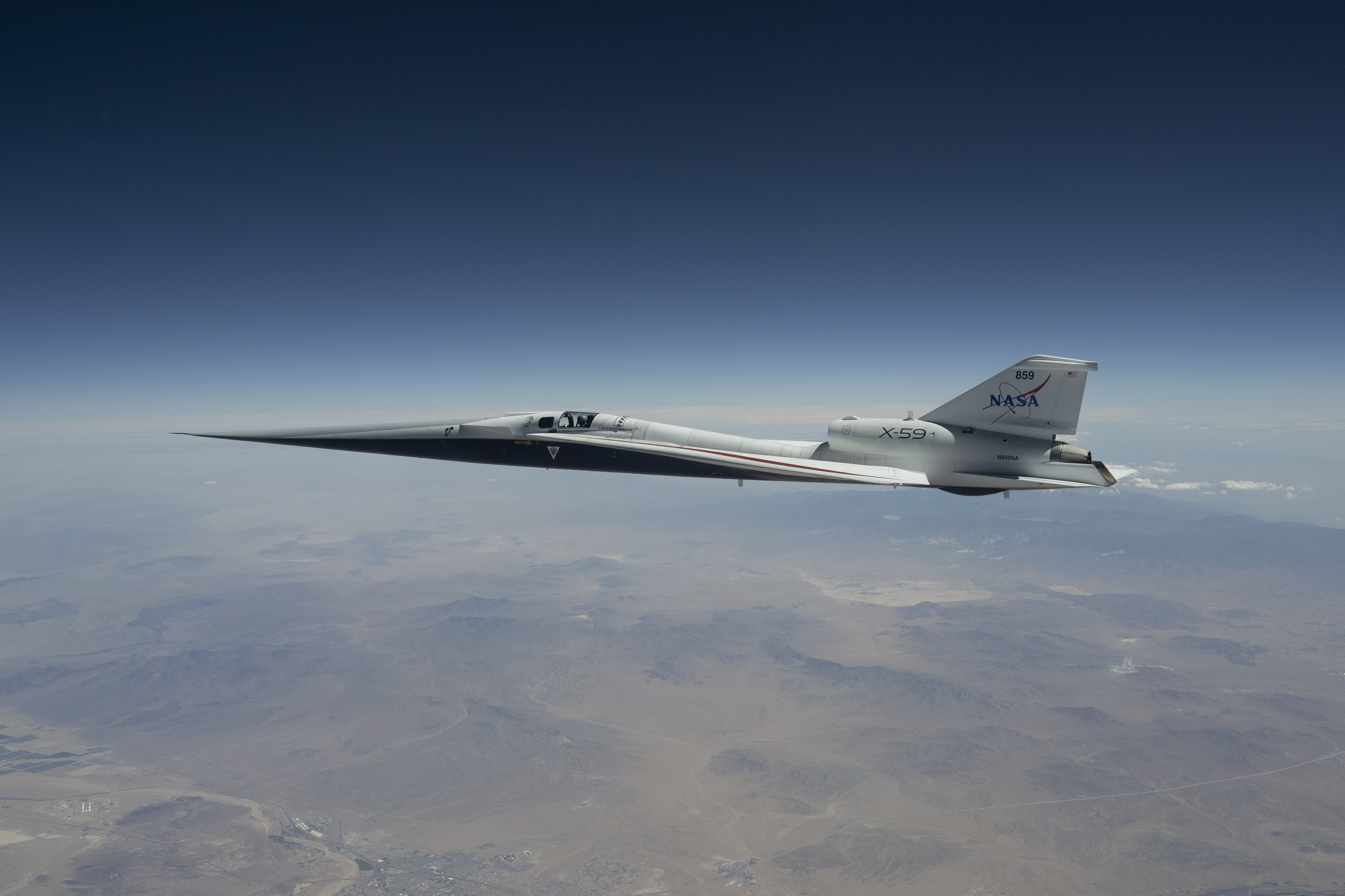 NASA’s X-59 quiet supersonic research aircraft flies high above the Mojave Desert. The aircraft is shown from a side profile with its long nose, swept wings and white fuselage stand out against the background.