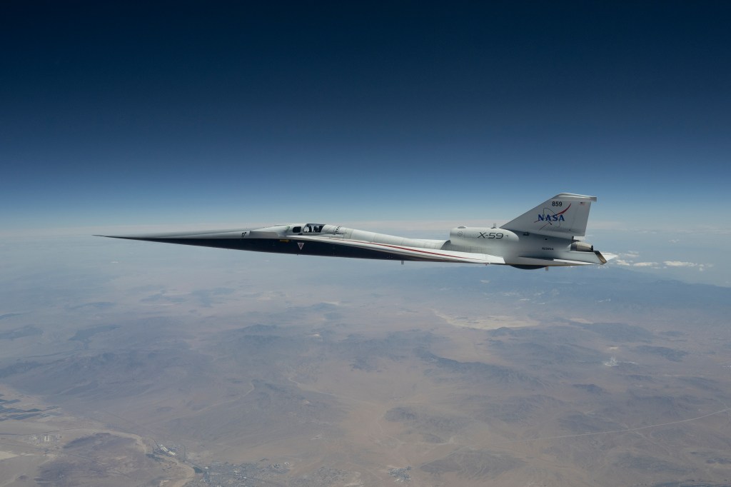 NASA’s X-59 quiet supersonic research aircraft flies high above the Mojave Desert. The aircraft is shown from a side profile with its long nose, swept wings and white fuselage stand out against the background.
