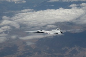 NASA’s X-59 quiet supersonic research aircraft flies high above the Mojave Desert. The aircraft is shown from a side profile with its long nose, swept wings and white fuselage stand out against the background.