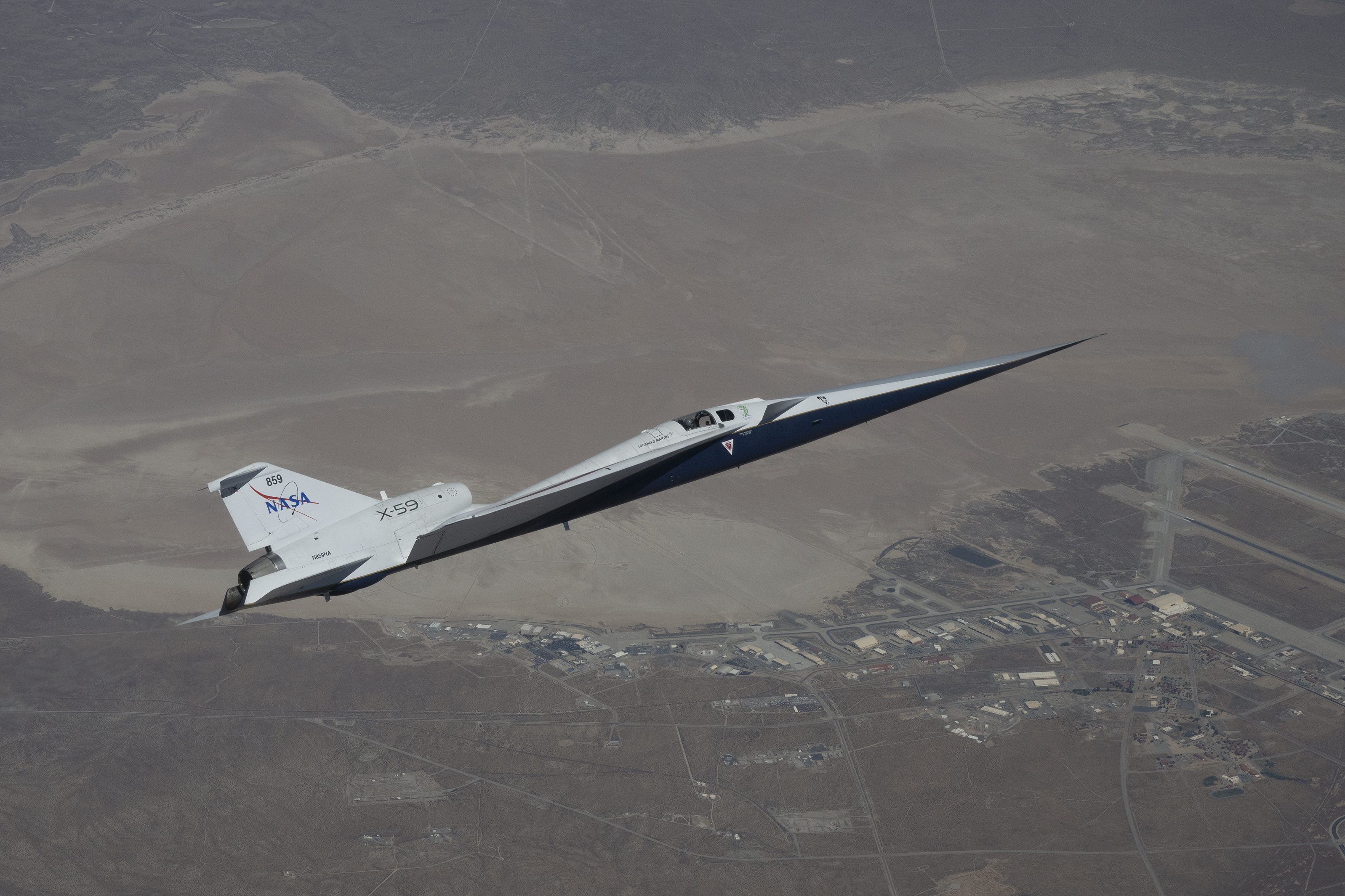 NASA’s X-59 quiet supersonic research aircraft flies high above the Mojave Desert. The aircraft is shown from a side profile with its long nose, swept wings and white fuselage stand out against the background.